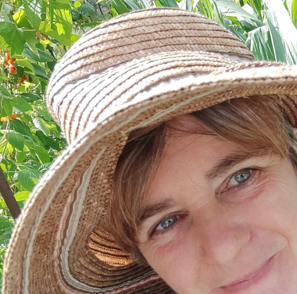 Photograph of a woman's face in a straw hat against a green background in a circle. The photo is cropped to show just the woman's eyes, nose and hat